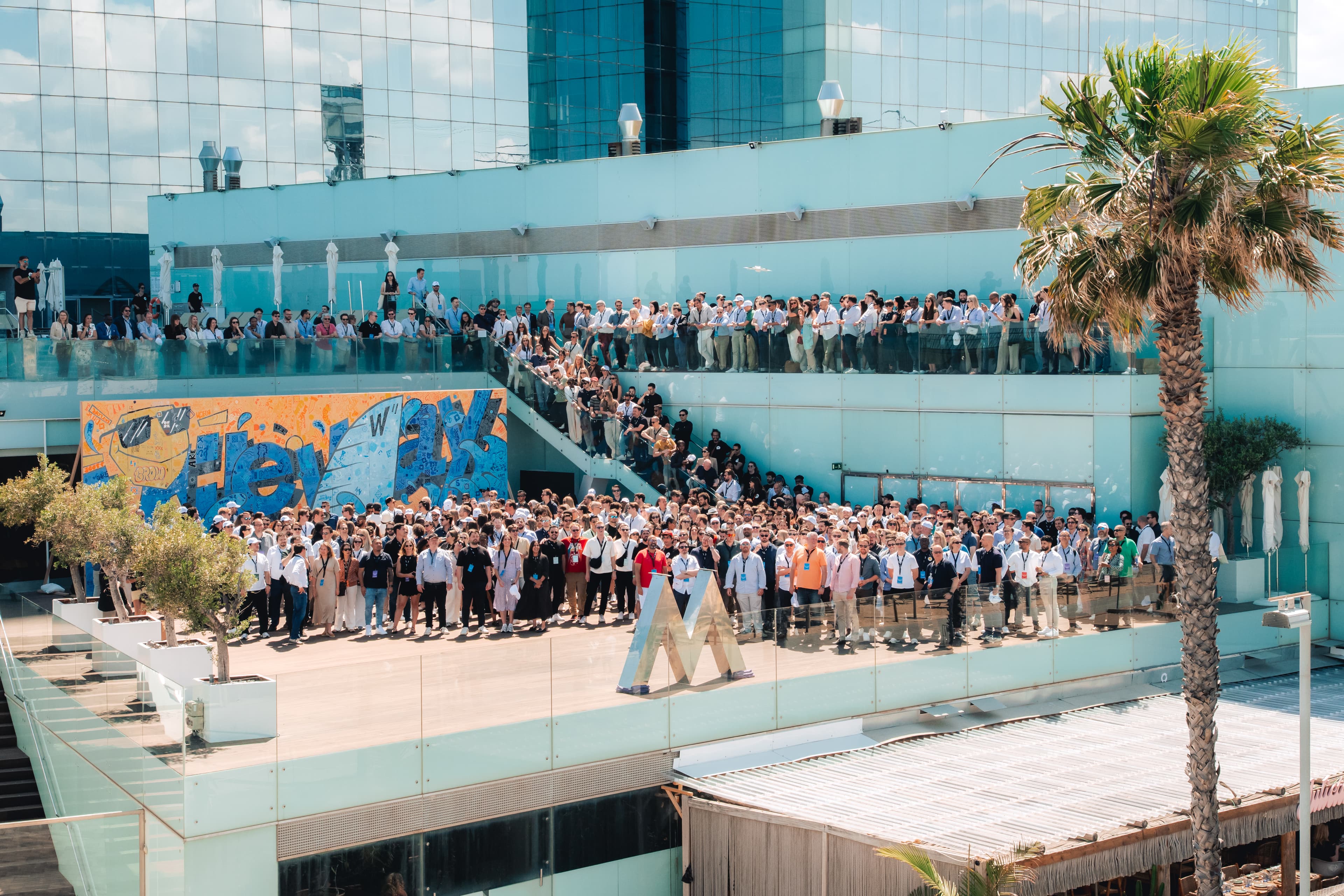A large group of people pose for a photo on multiple levels of an outdoor terrace by a modern glass building with a mural and palm tree visible.