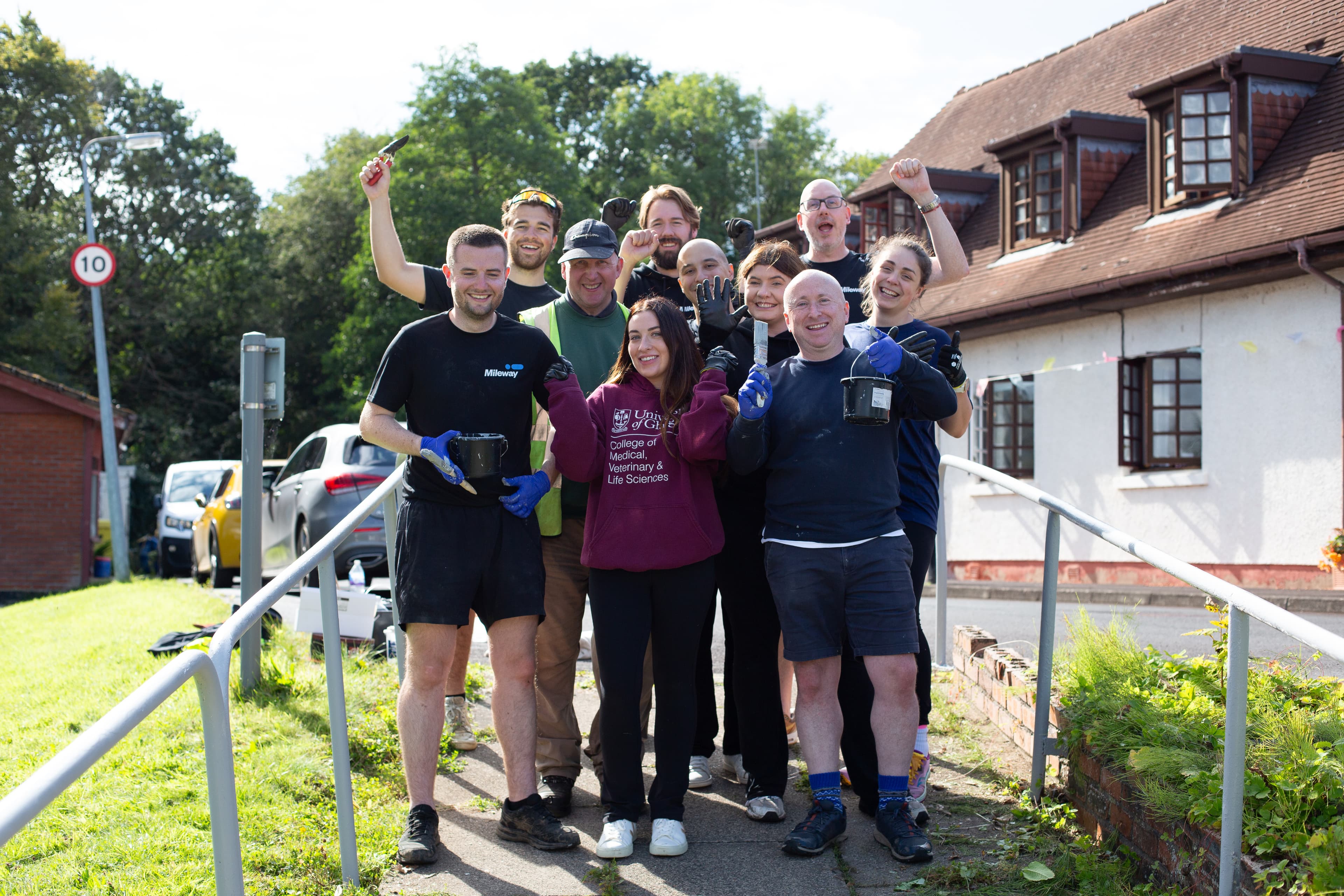 A group of people stand outdoors by a building, smiling and holding painting supplies, appearing to be part of a volunteer or community project.