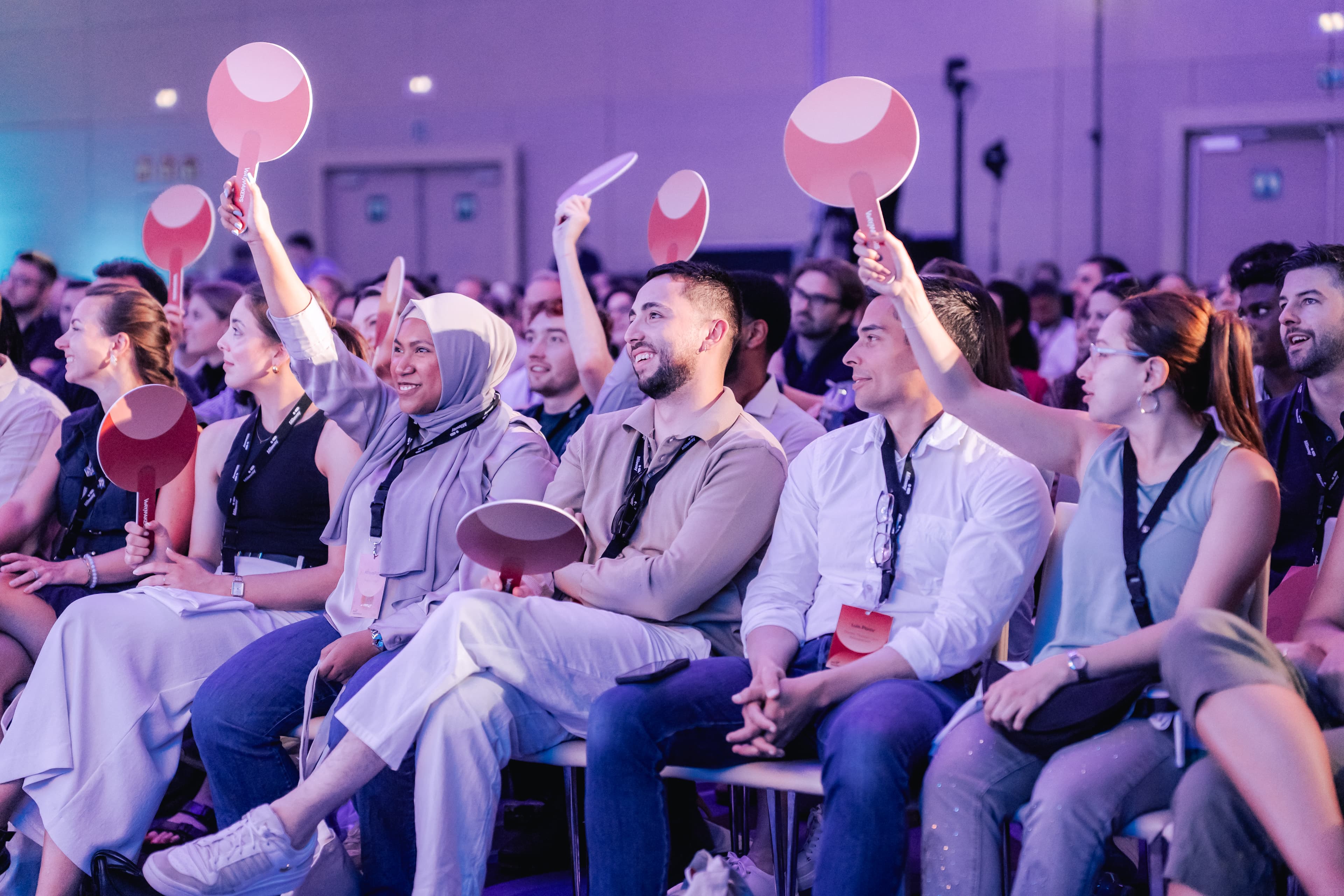 A group of people seated in rows at an event, smiling and holding up circular paddles, with lanyards round their necks in a brightly lit room.