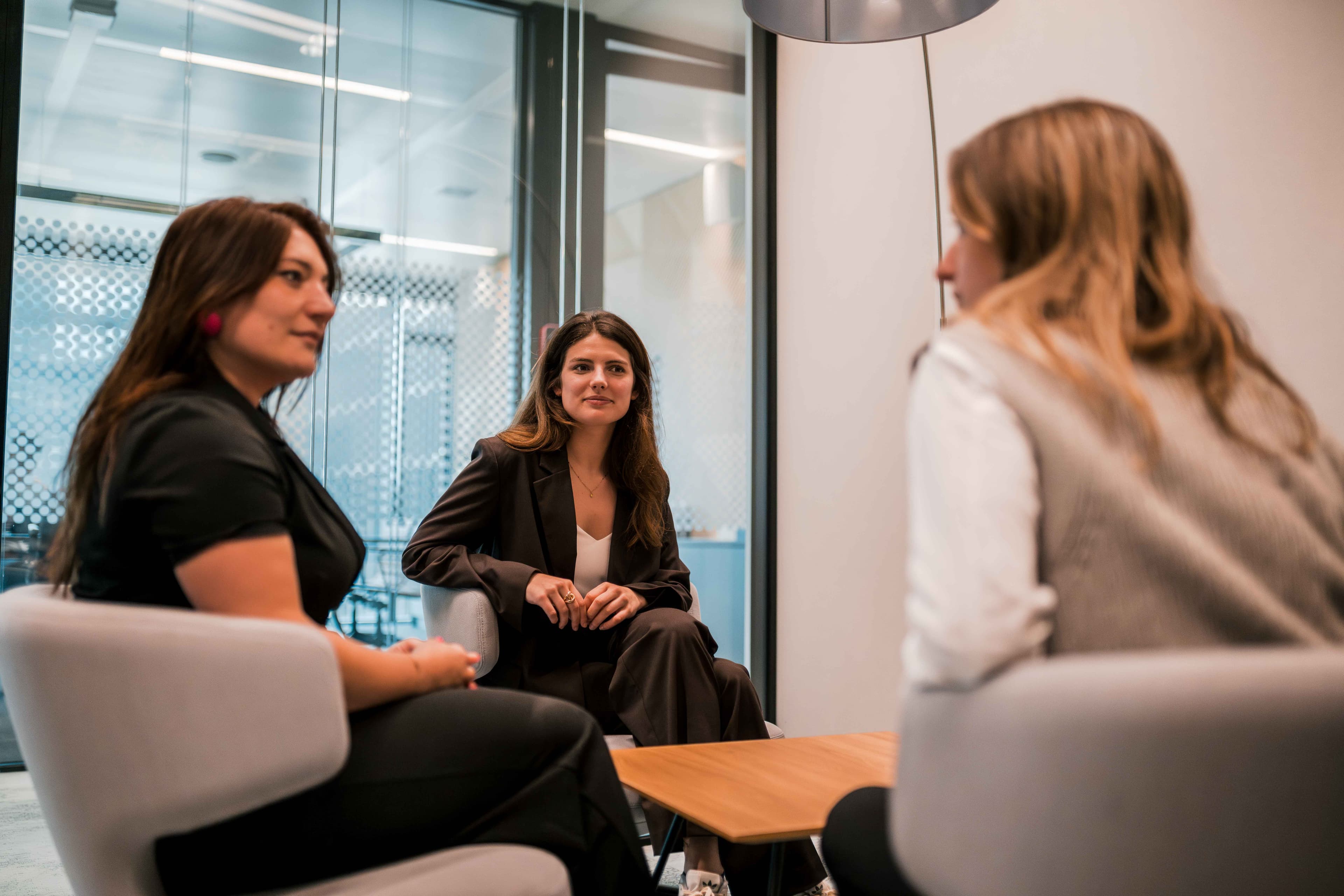 Three women sit in modern office chairs around a small wooden table, engaged in conversation inside a glass-walled meeting room.