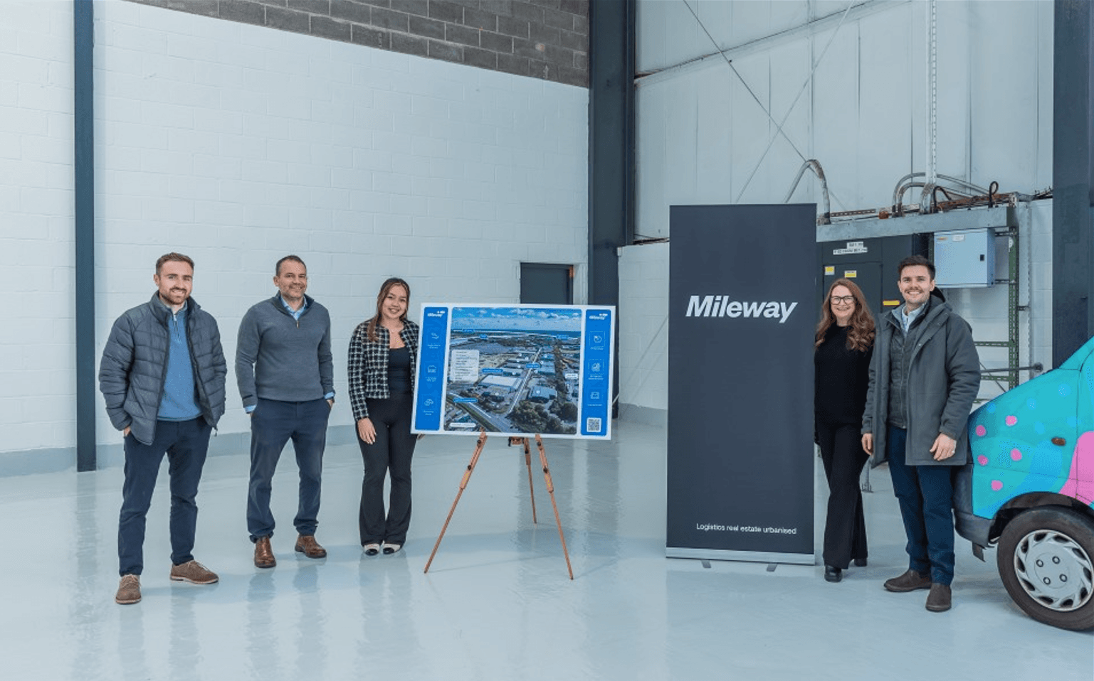 Five people stand indoors next to a presentation board with an aerial site image and a “Mileway” banner in an industrial warehouse setting.