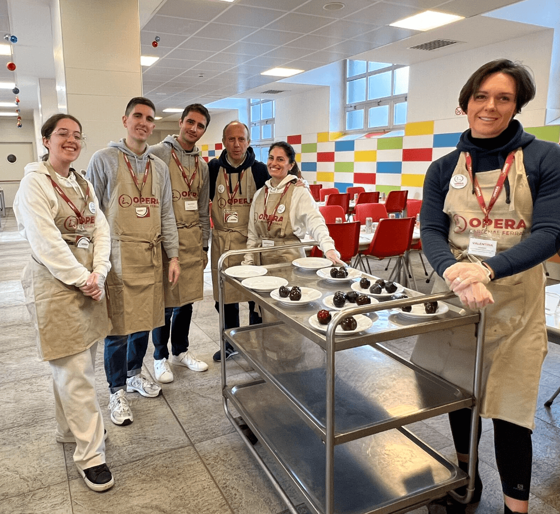 Six people wearing aprons stand around a food trolley with puddings on plates in a brightly lit canteen with red chairs and a colourful tiled wall.