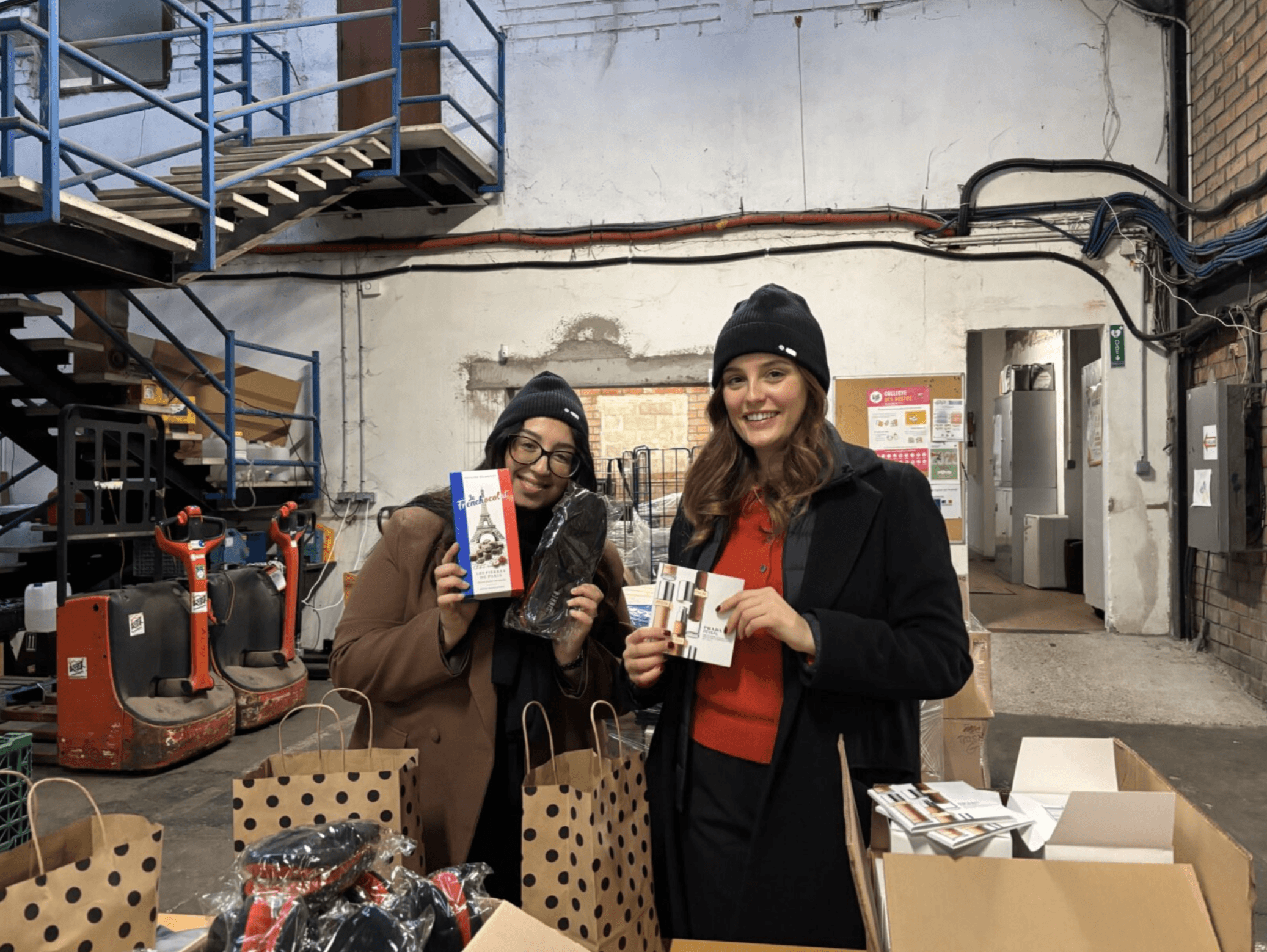 Two people wearing winter coats and beanies are standing indoors, holding gift items and packing paper bags on a table in an industrial setting.