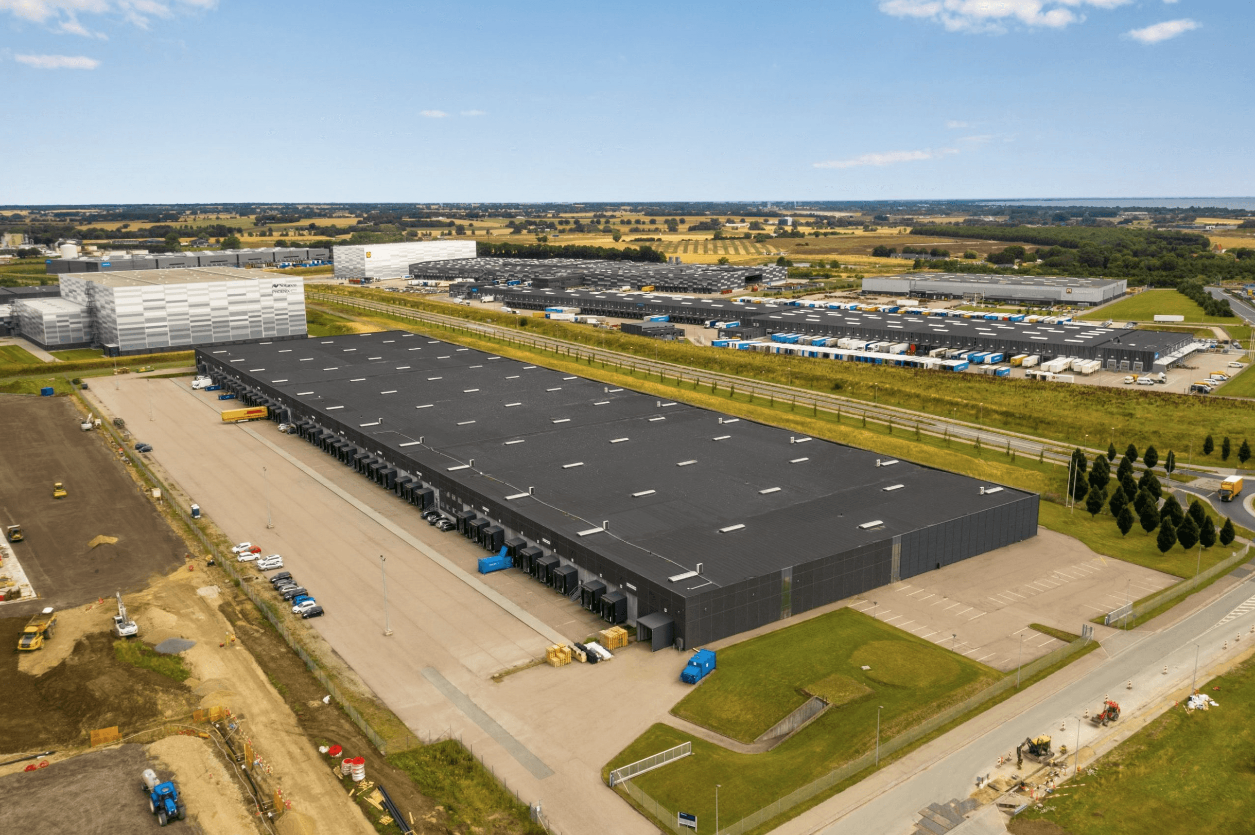 Aerial view of a large industrial warehouse with loading bays, surrounded by car parks, lorries, and fields under a partly cloudy sky.