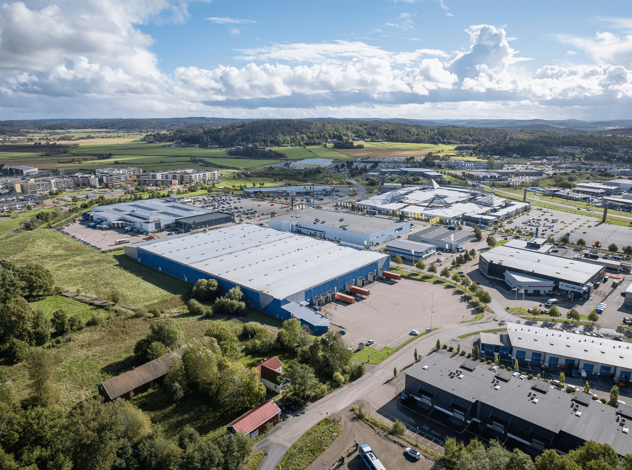 Aerial view of a large industrial area with multiple warehouses, car parks, and green fields surrounding the buildings under a partly cloudy sky.