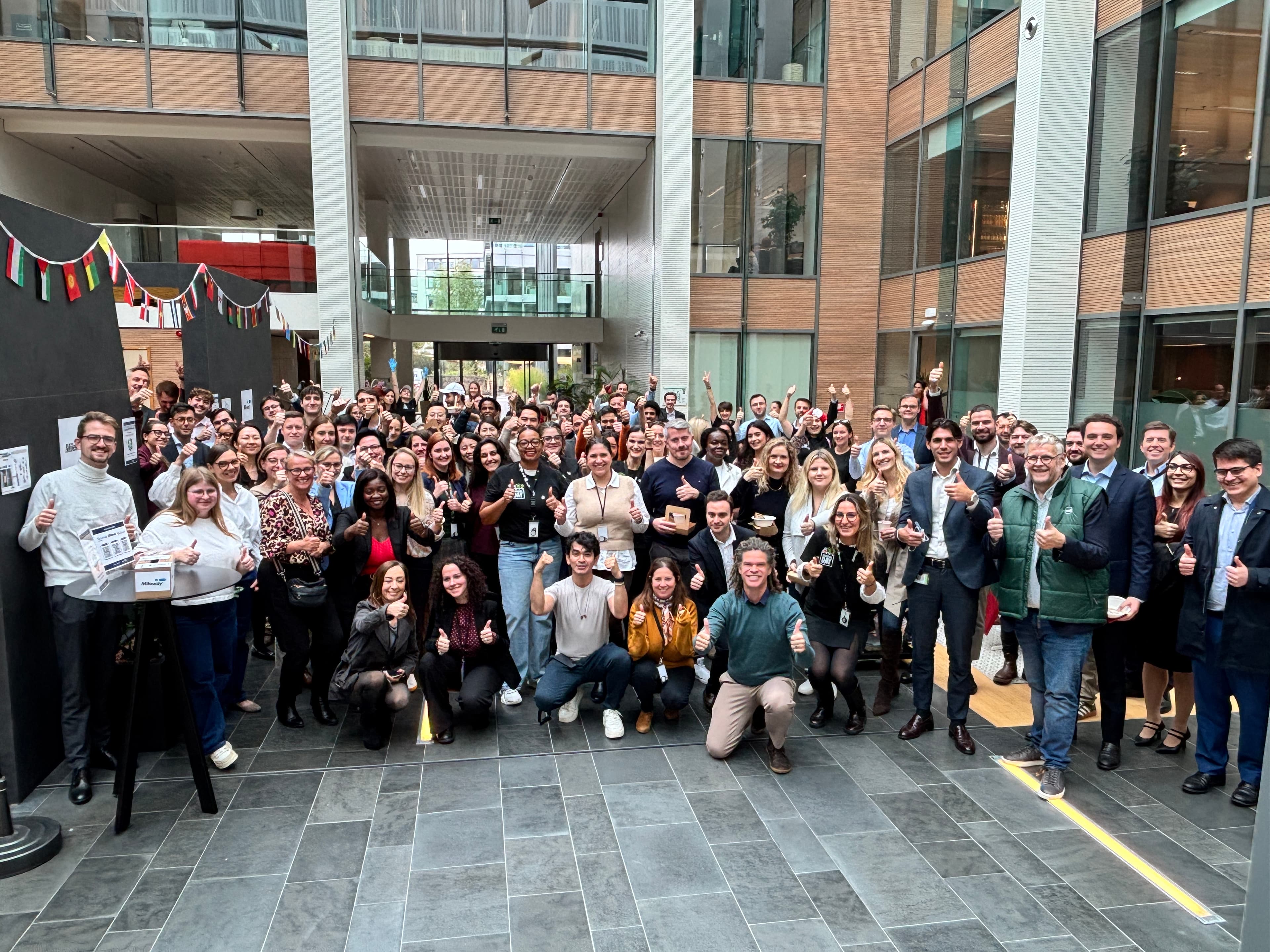 A large group of people standing indoors in an atrium, smiling and giving thumbs up whilst facing the camera.
