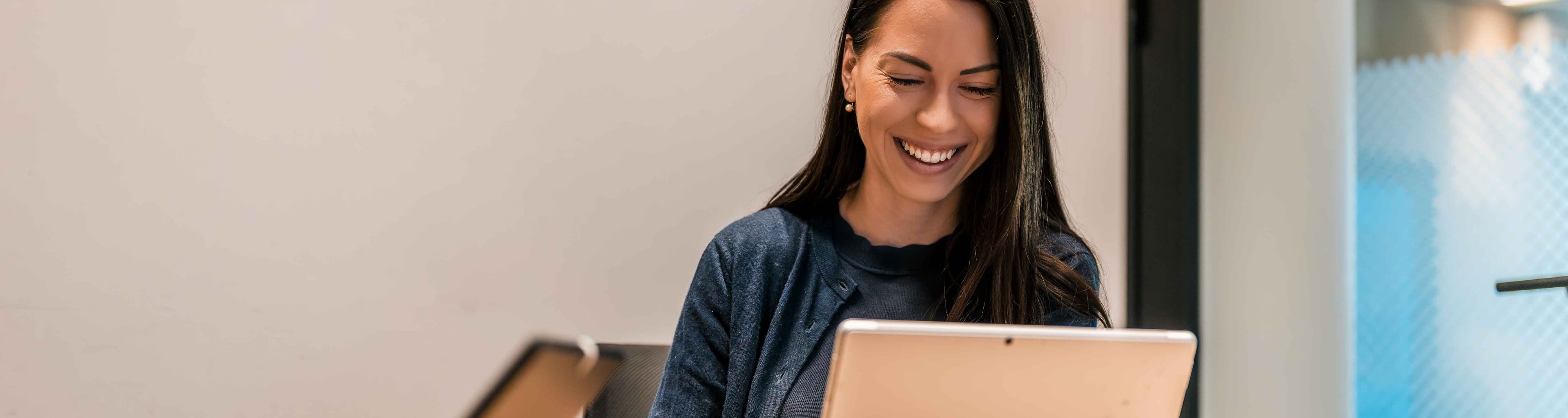 A person sits at a table indoors, smiling while looking at a tablet device.