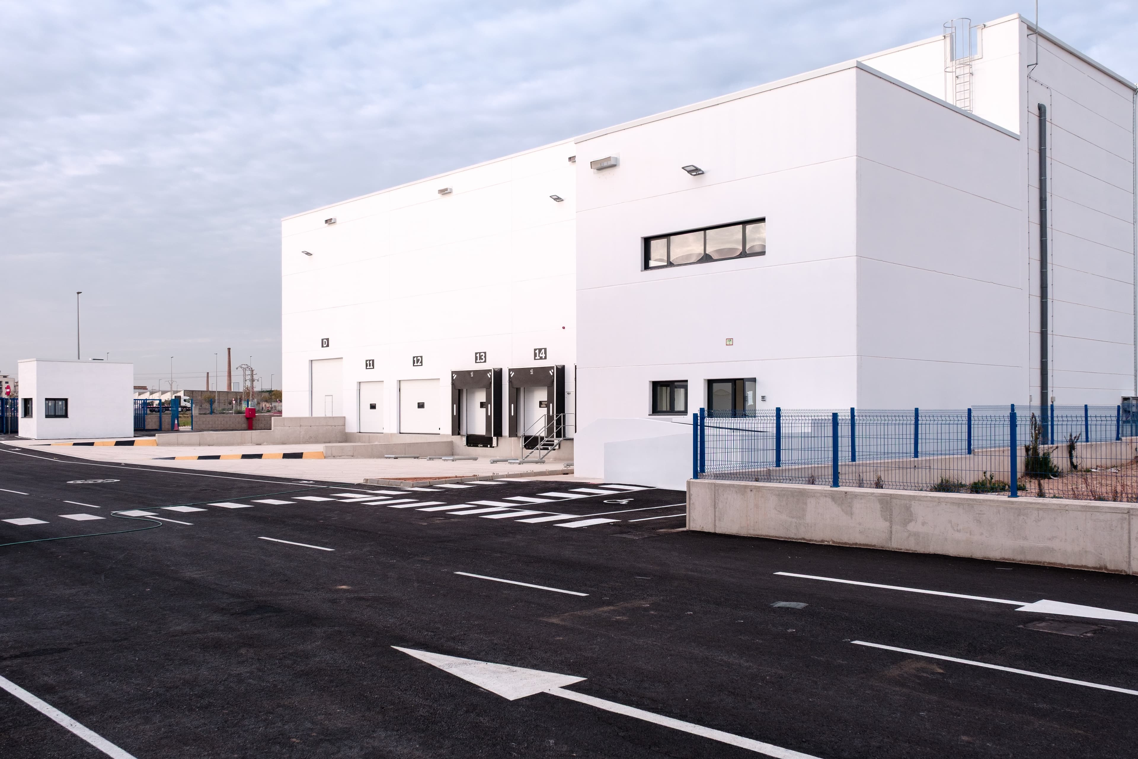 A modern white industrial warehouse with multiple loading docks, blue fencing, and an empty asphalt parking area under a cloudy sky.