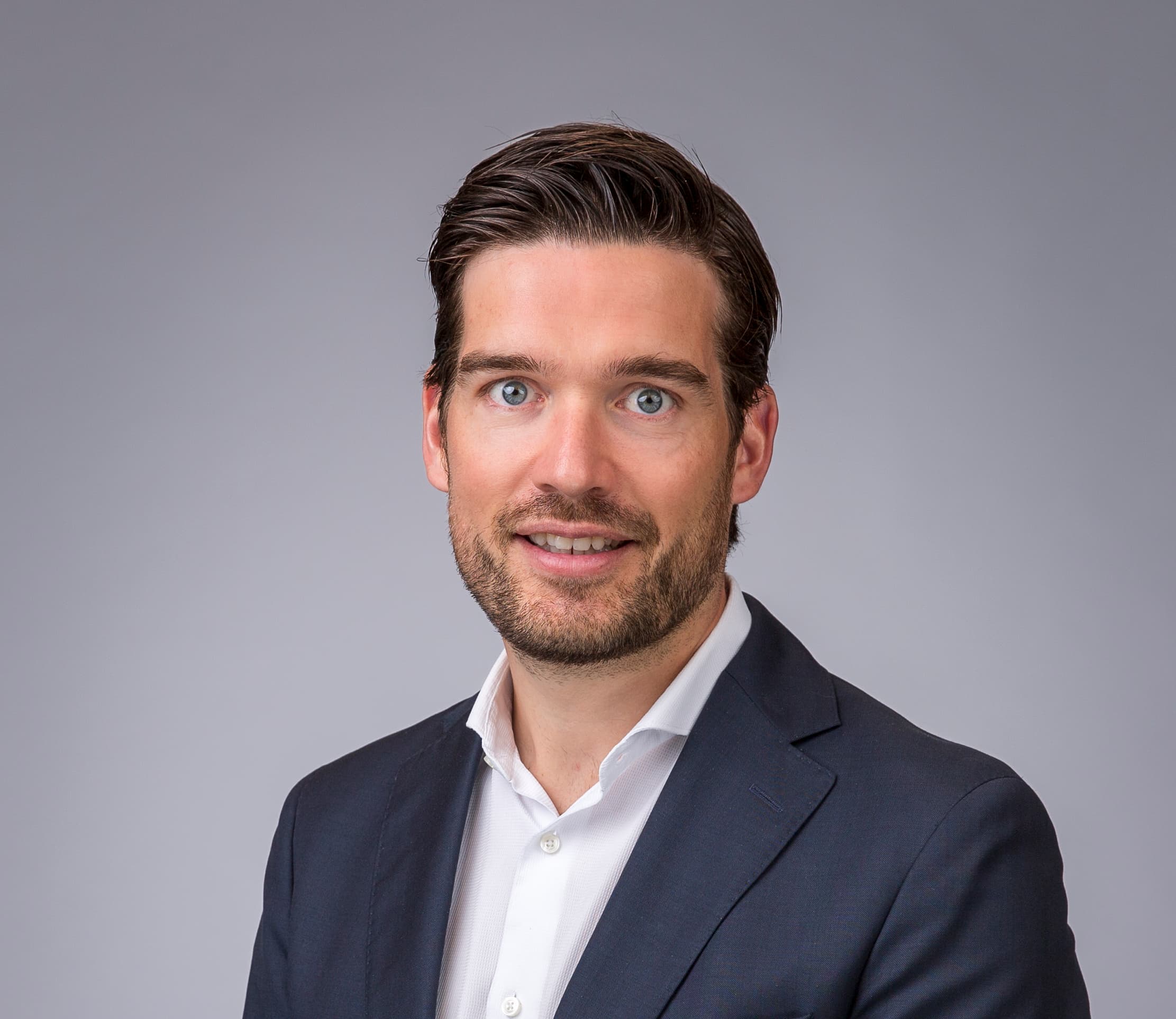 A person in a dark suit and white shirt stands against a plain grey background, facing the camera.