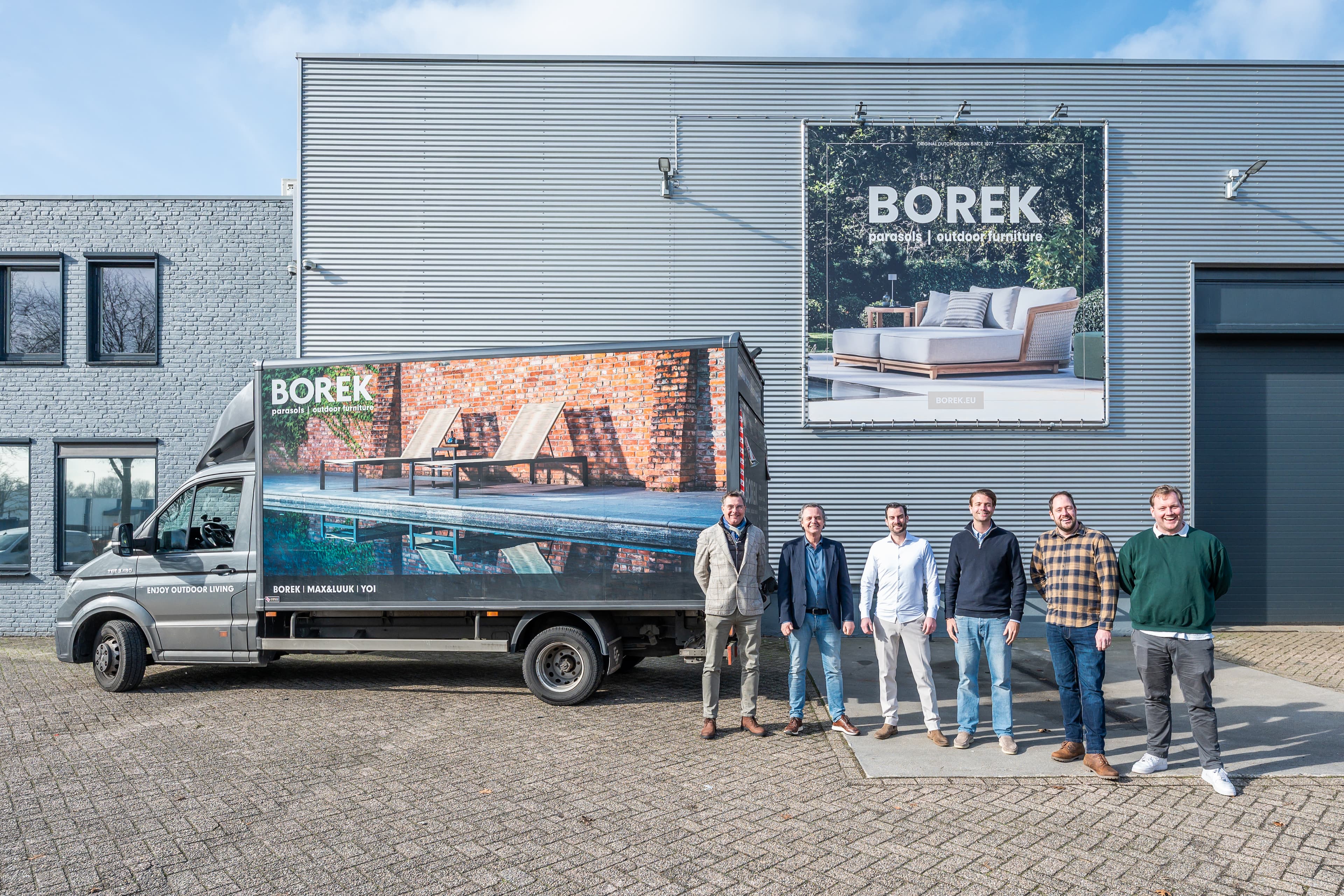 A group stands in front of a building with a BOREK sign, beside a van displaying an outdoor furniture advert.