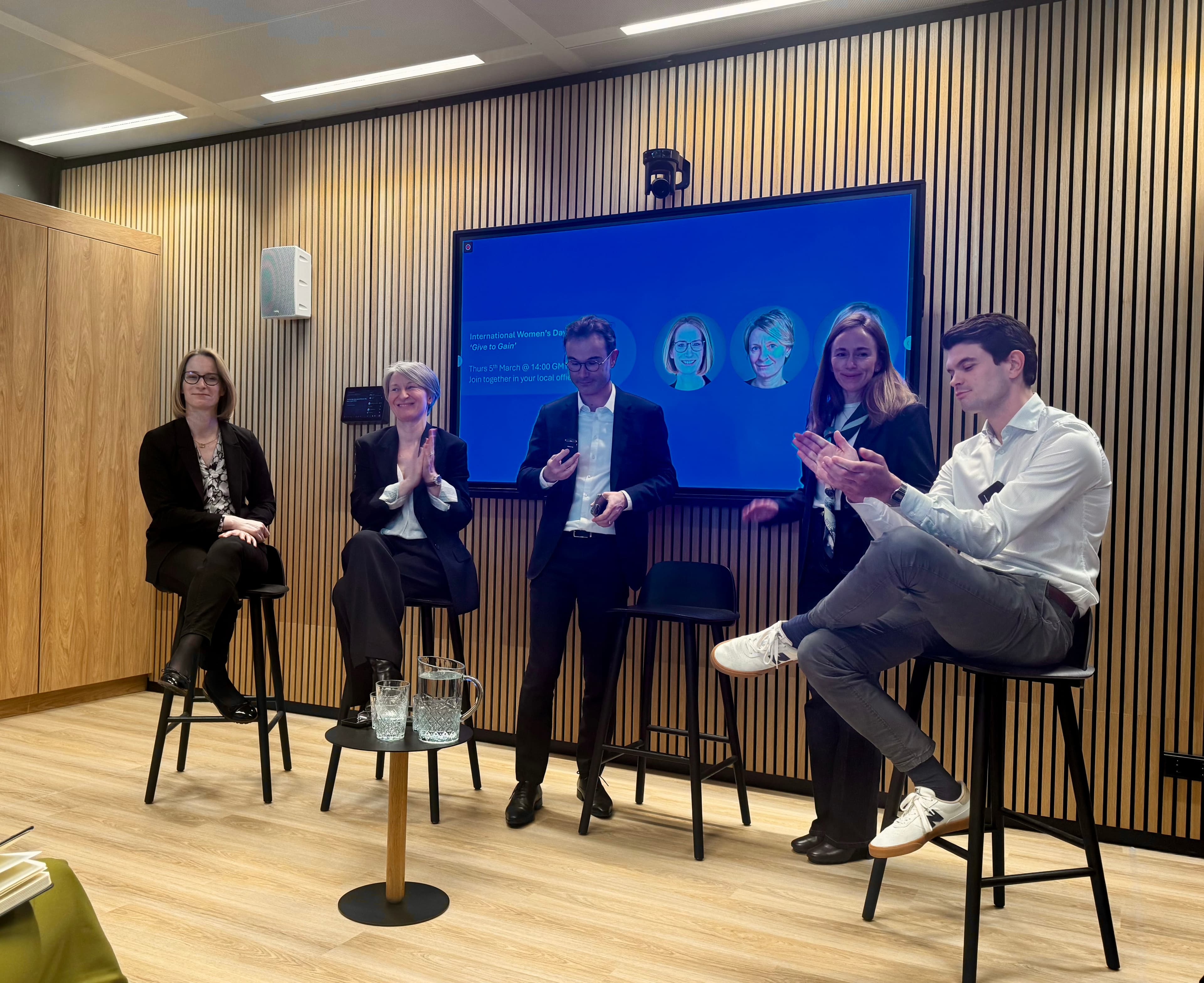 Several people sit and stand on a stage discussing IWD 2026's theme, Give to Gain.