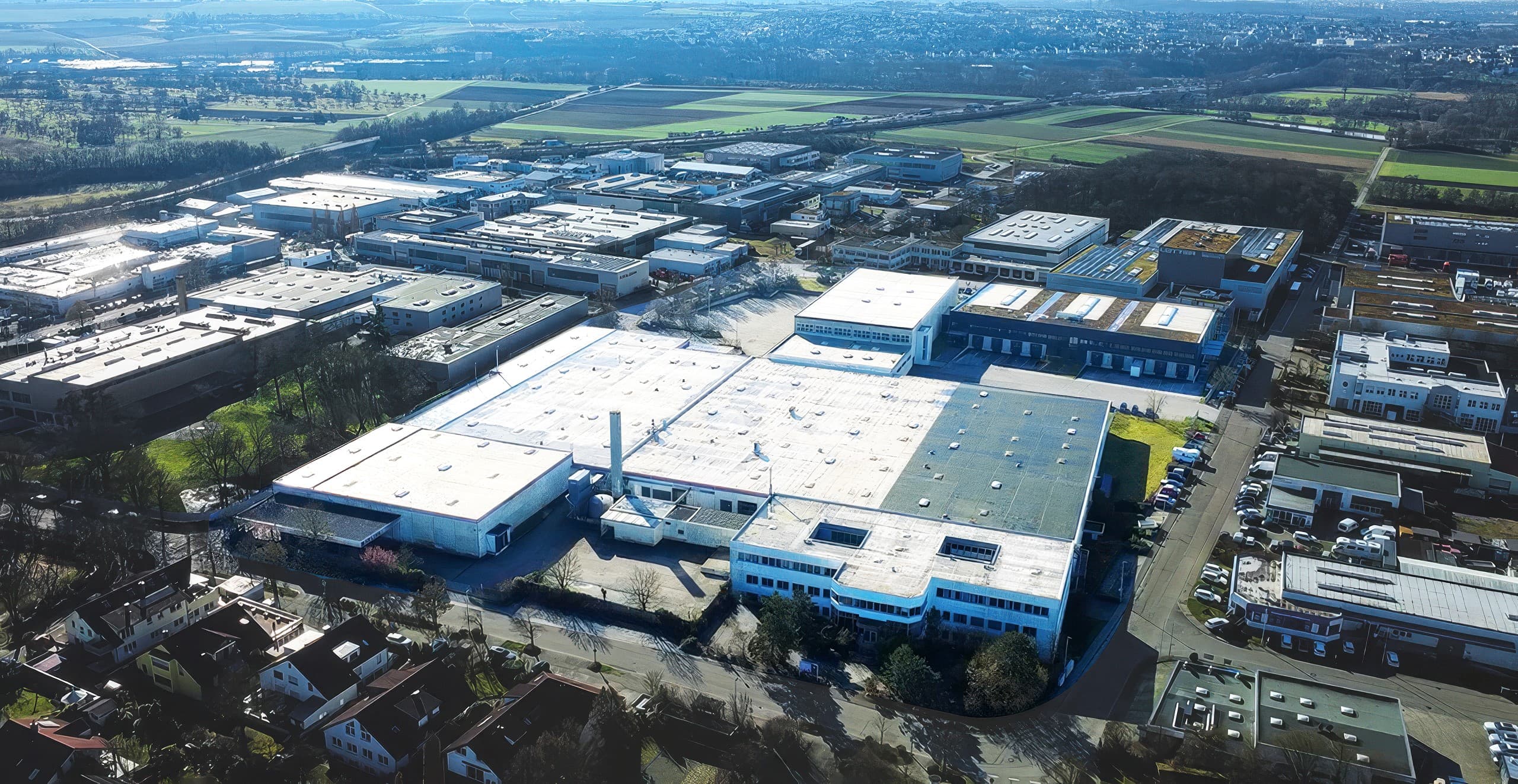 Aerial view of an industrial estate with warehouses, roads, and car parks, bordered by fields and distant residential areas.