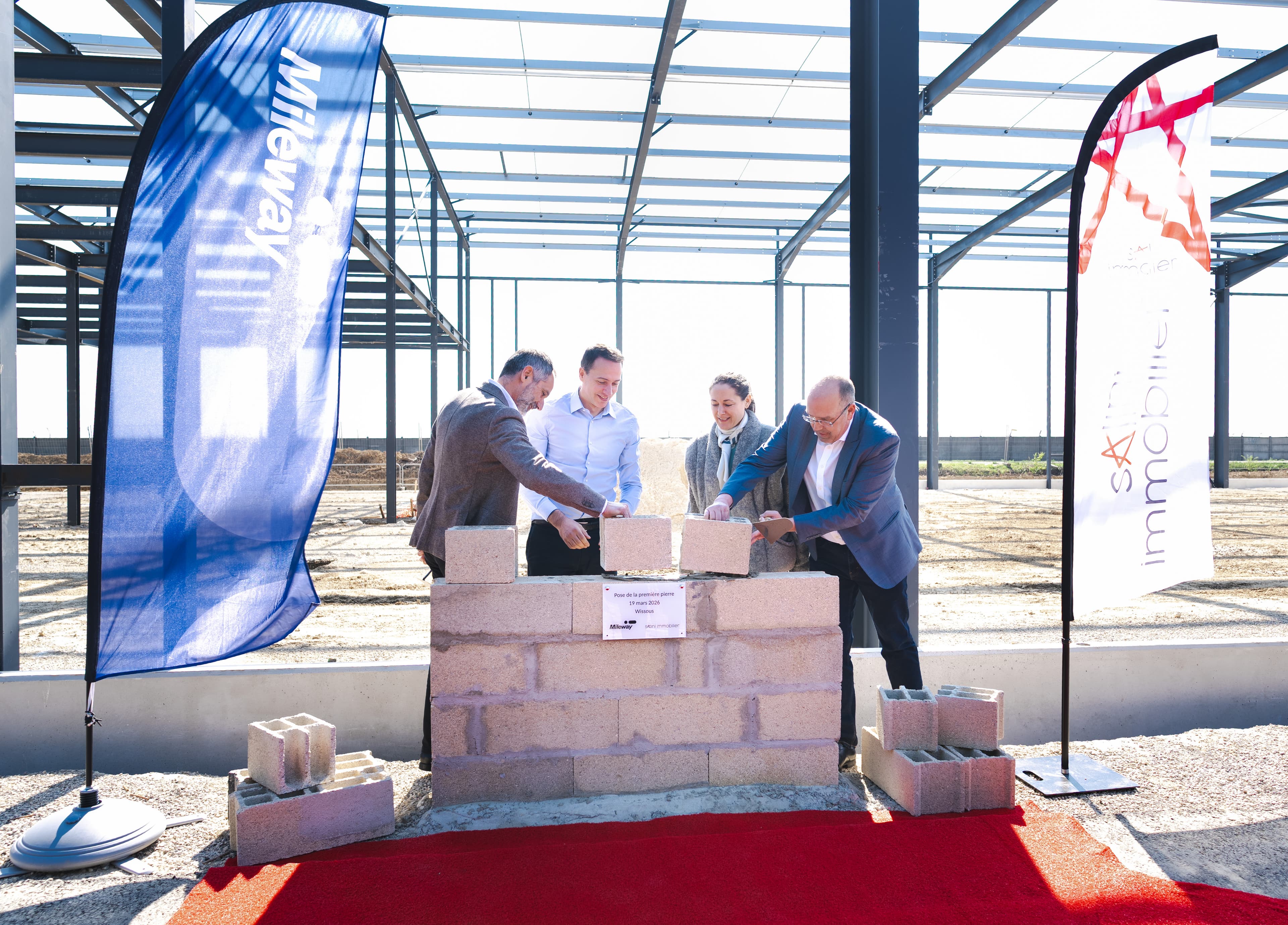 People lay bricks at a ceremonial event, with two flags and a building frame on a construction site in the background.