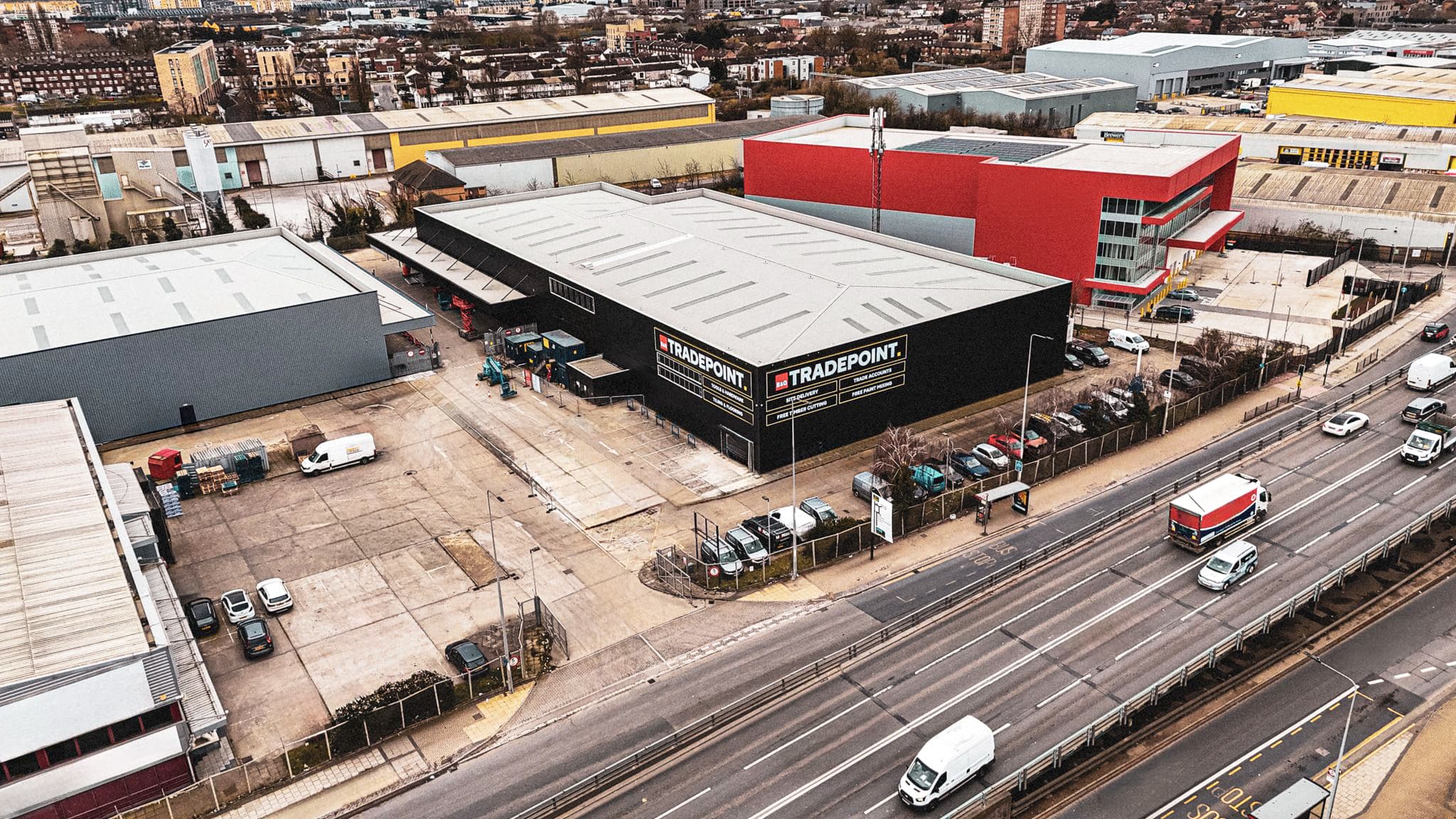 Aerial view of an industrial estate featuring a TradePoint warehouse, car park, delivery vehicles, and nearby roads with traffic.