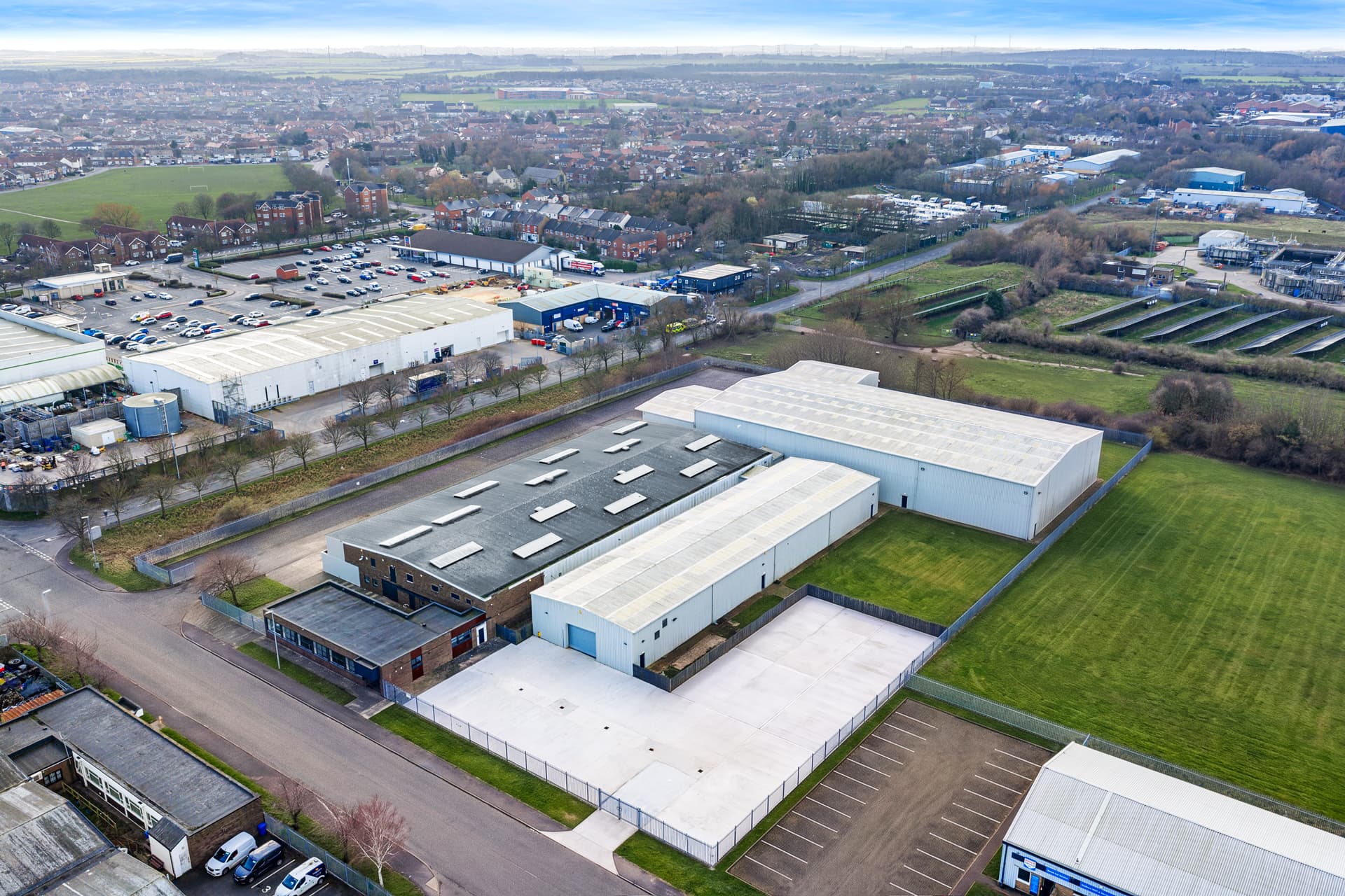 Aerial view of an industrial estate featuring large warehouses, car parks, and green fields on the outskirts of a town.