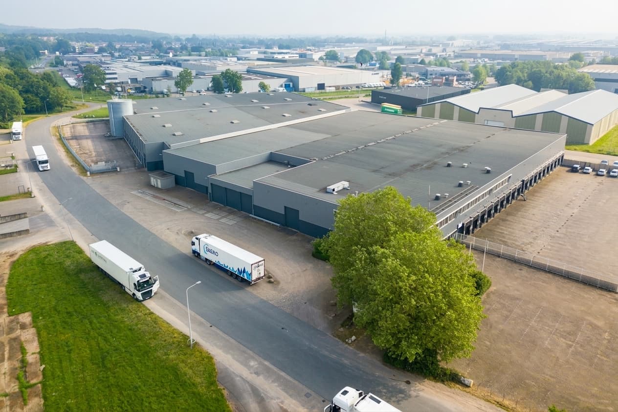 Aerial view of a large warehouse with loading bays, lorries parked nearby, trees, and surrounding industrial buildings.